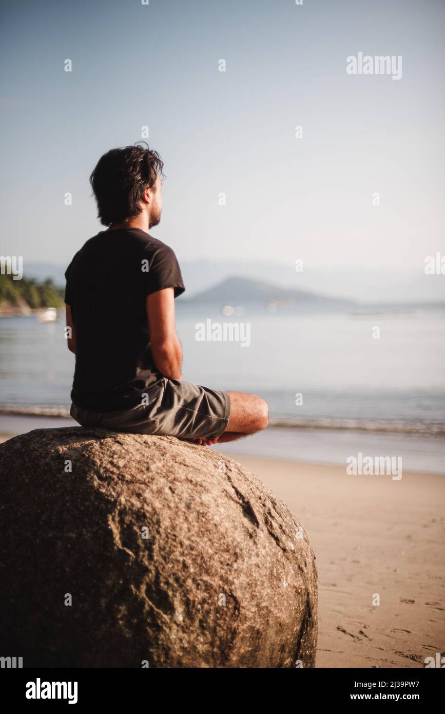 Uomo che si sieta su una roccia meditando sulla spiaggia Foto Stock