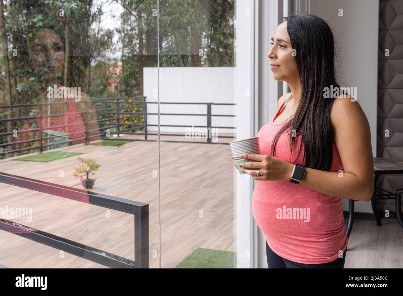 Giovane donna incinta con lunghi capelli neri lisci che guardano fuori la finestra della sua casa in una terrazza mentre si beve e la sua immagine si riflette Foto Stock