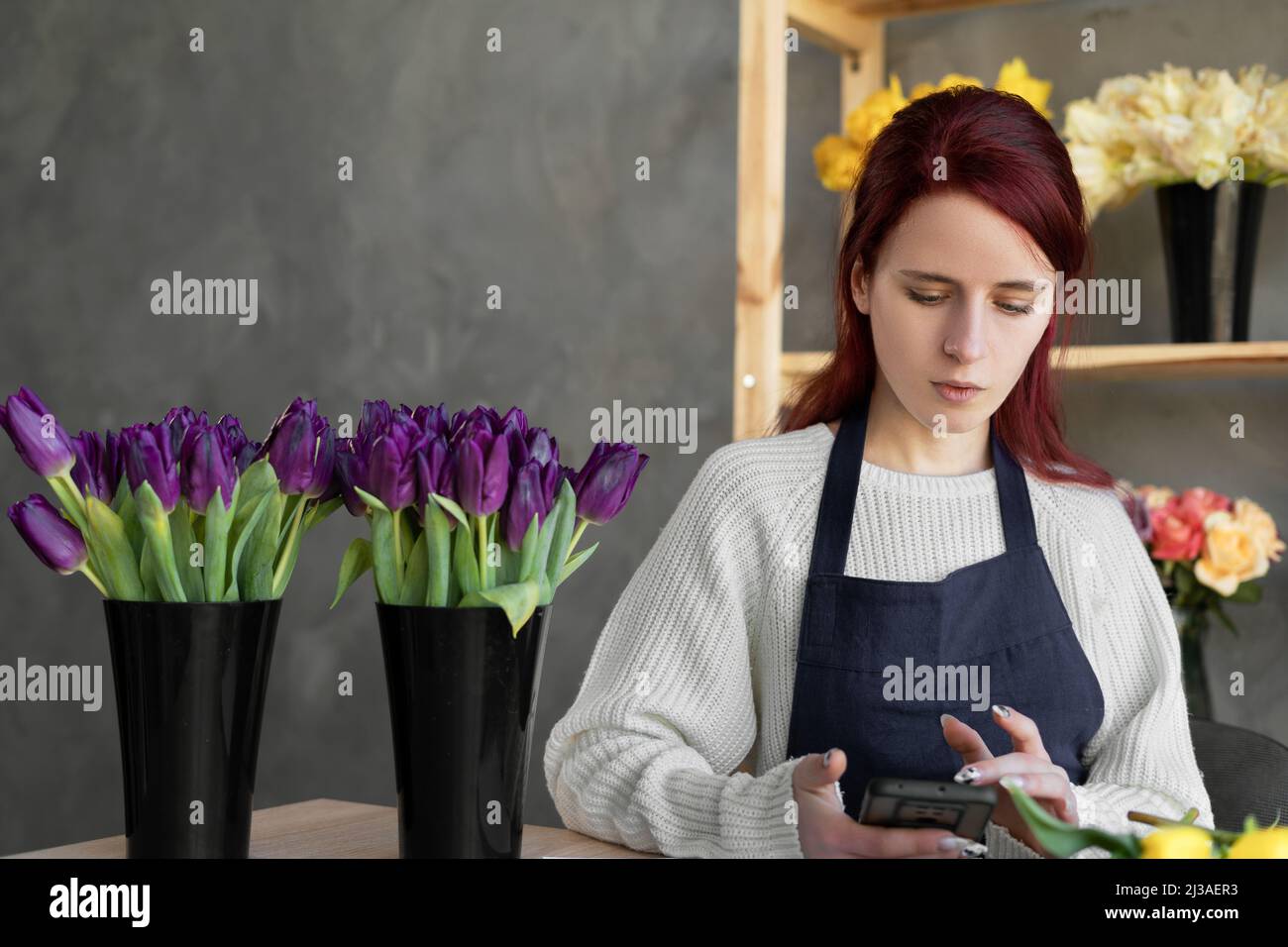 Un fiorista sorridente in un grembiule blu controlla gli ordini sul sito utilizzando app e un telefono cellulare mentre si siede a un tavolo con fiori freschi. Il concetto di Foto Stock