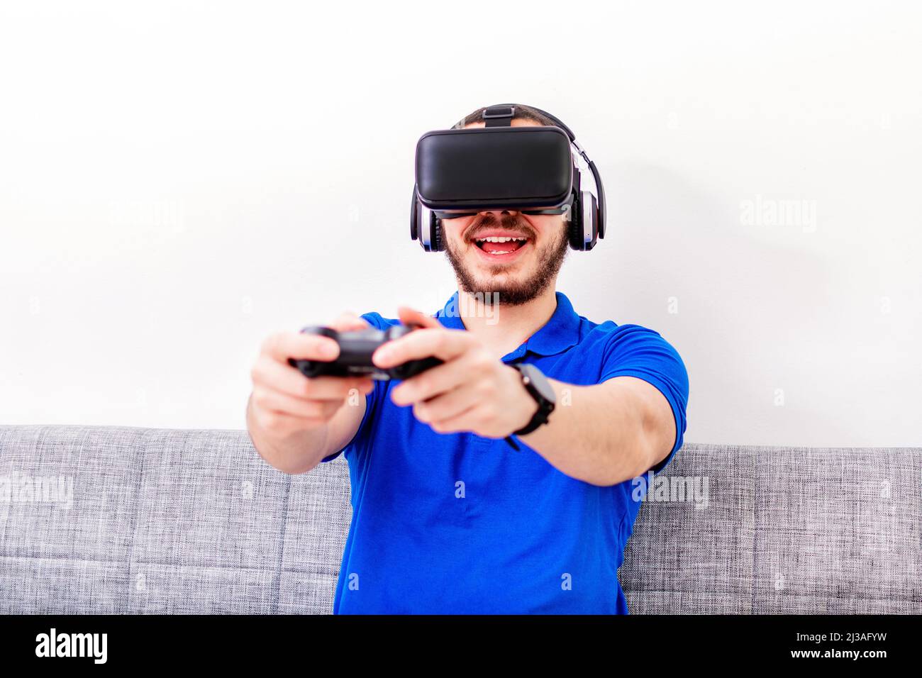 Un uomo bearded medio orientale seduto sul divano usando la cuffia e il controller di realtà virtuale. Foto Stock