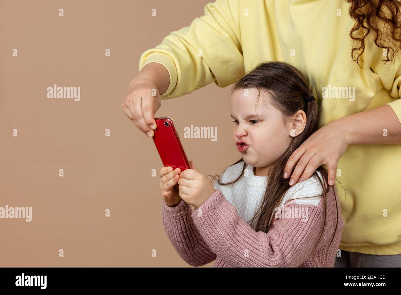 Ritratto del genitore che porta lo smartphone fuori dalle mani delle ragazze, sfondo beige. Figlia insoddisfatta tenere il telefono e combattere indietro. Concetto di prevenzione di Foto Stock