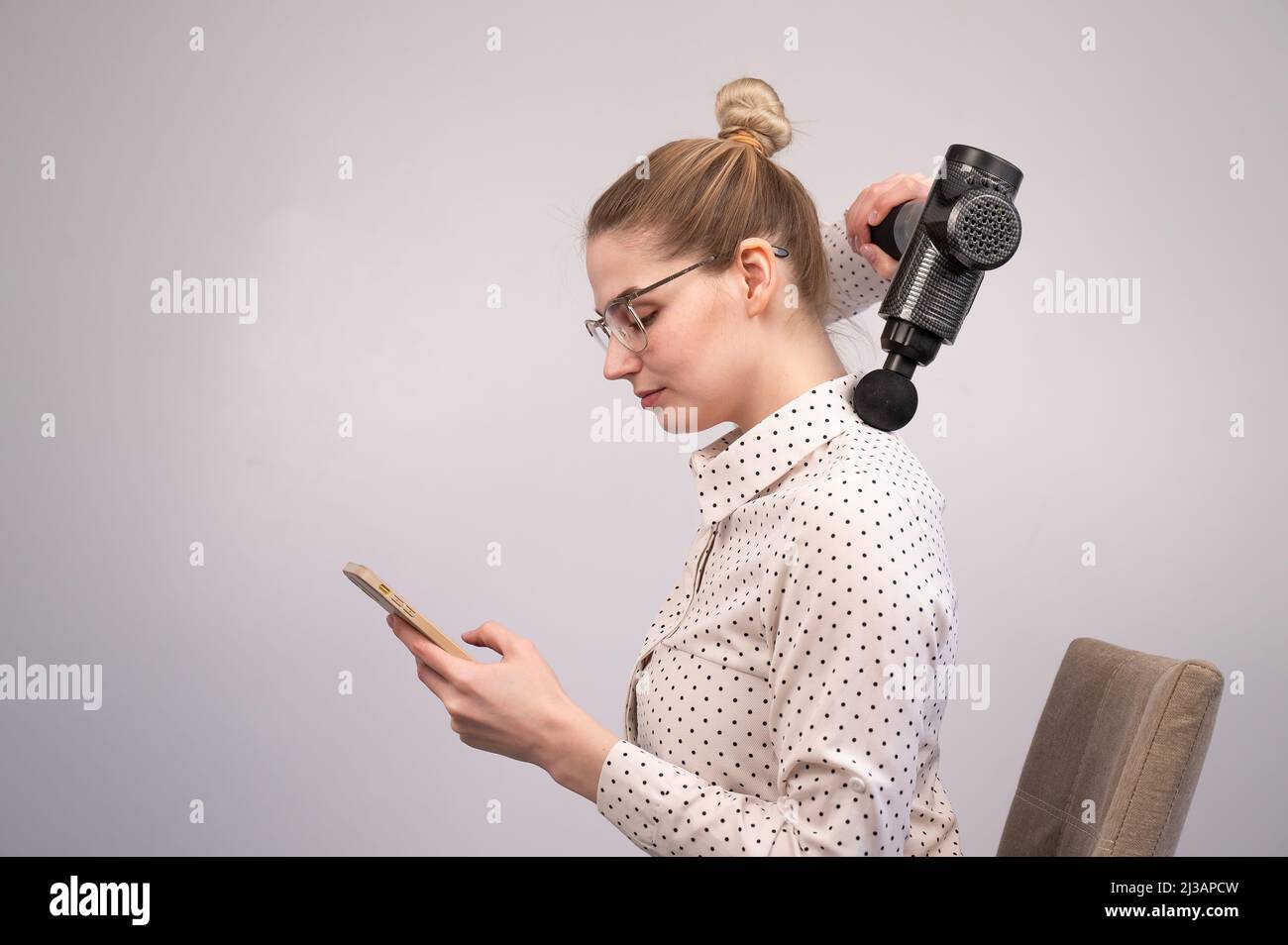 Una giovane donna si siede su una sedia, usa uno smartphone e si fa un massaggio con una pistola da massaggio elettro. Foto Stock