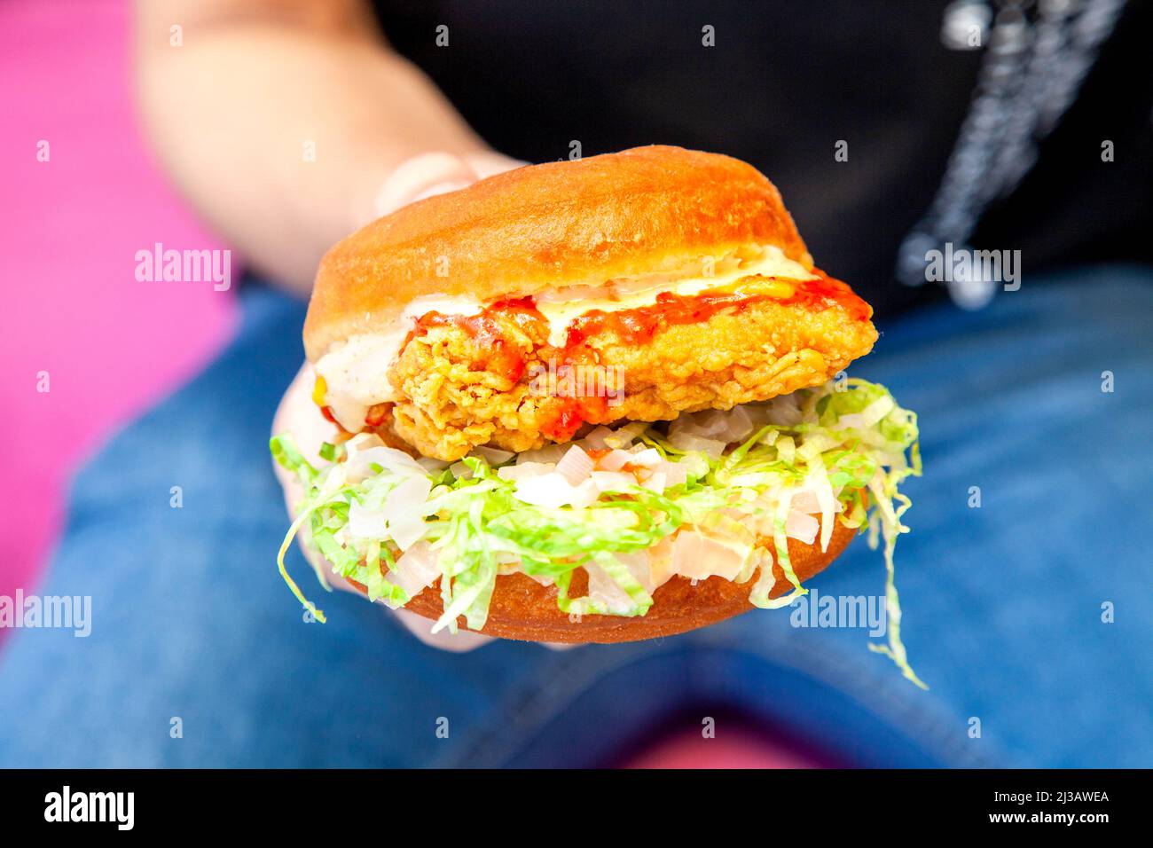 Hamburger di strada di pollo fritto succoso con una mano in primo piano con una persona che indossa jeans blu e camicia nera sullo sfondo Foto Stock
