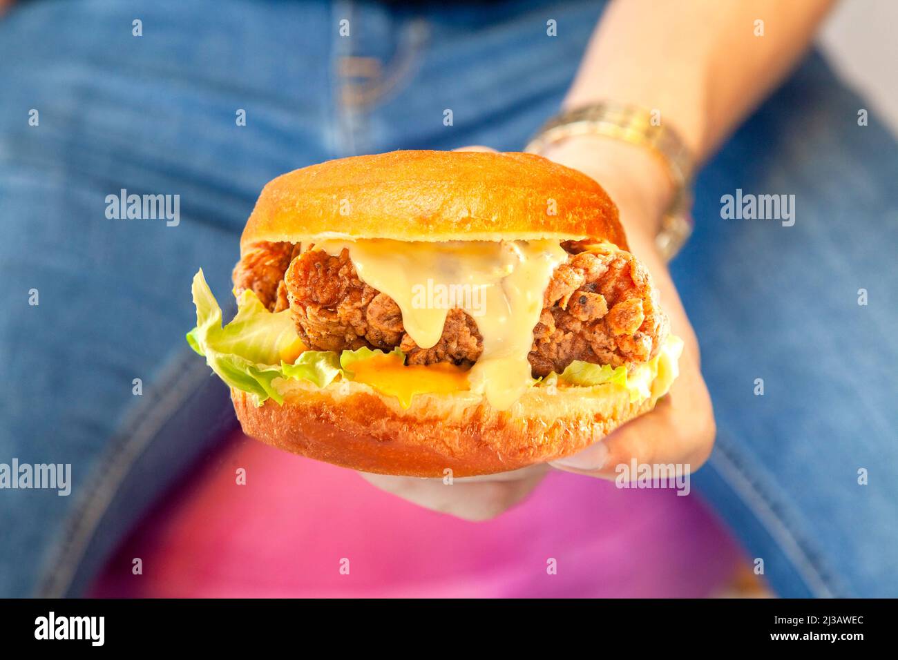 Hamburger di strada di pollo fritto succoso con una mano in primo piano con una persona che indossa jeans blu e camicia nera sullo sfondo Foto Stock