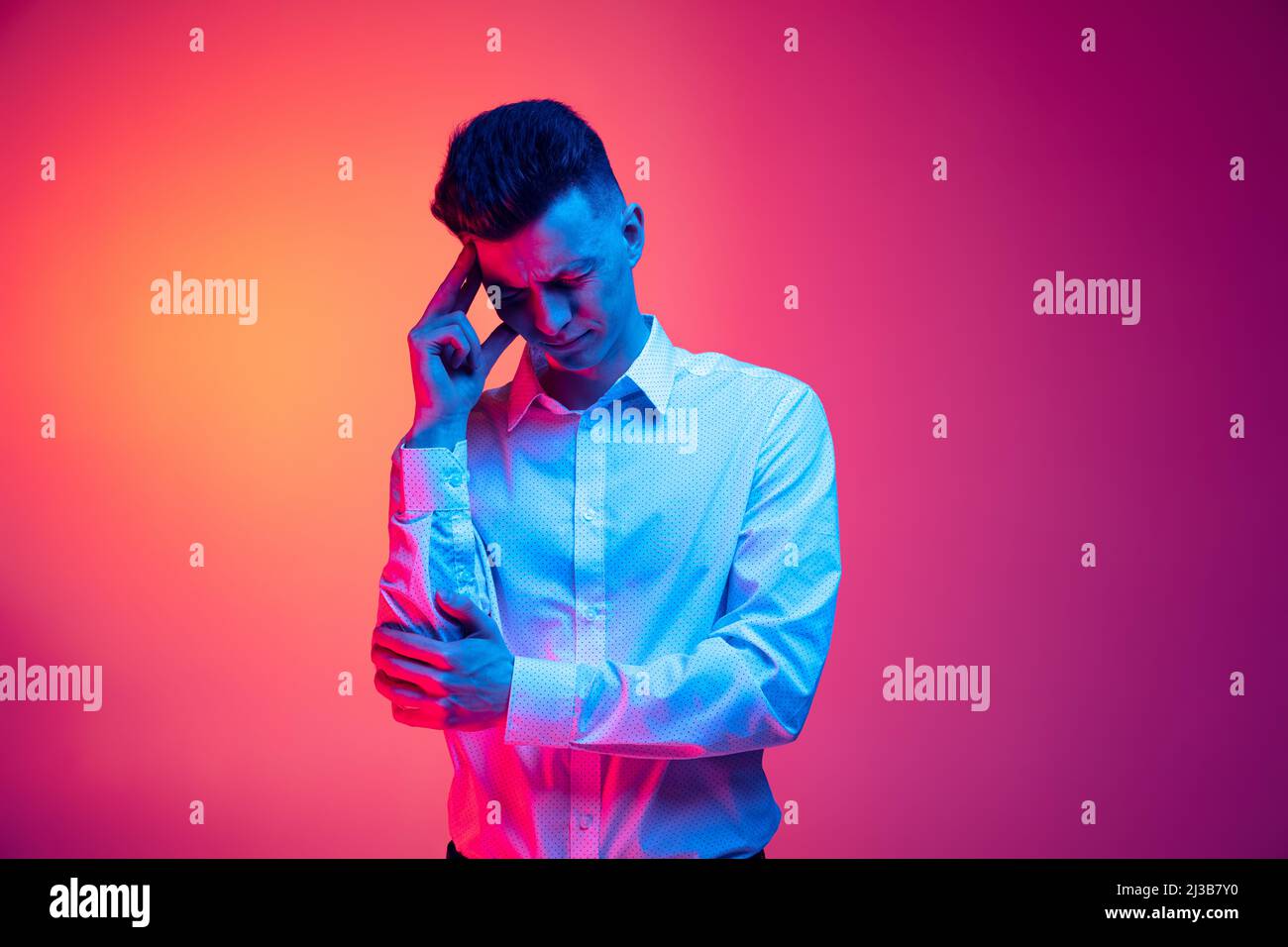 Ritratto a mezza lunghezza di giovane uomo bello in camicia bianca ha un terribile mal di testa isolato su sfondo sttudio color magenta in luce neon blu Foto Stock