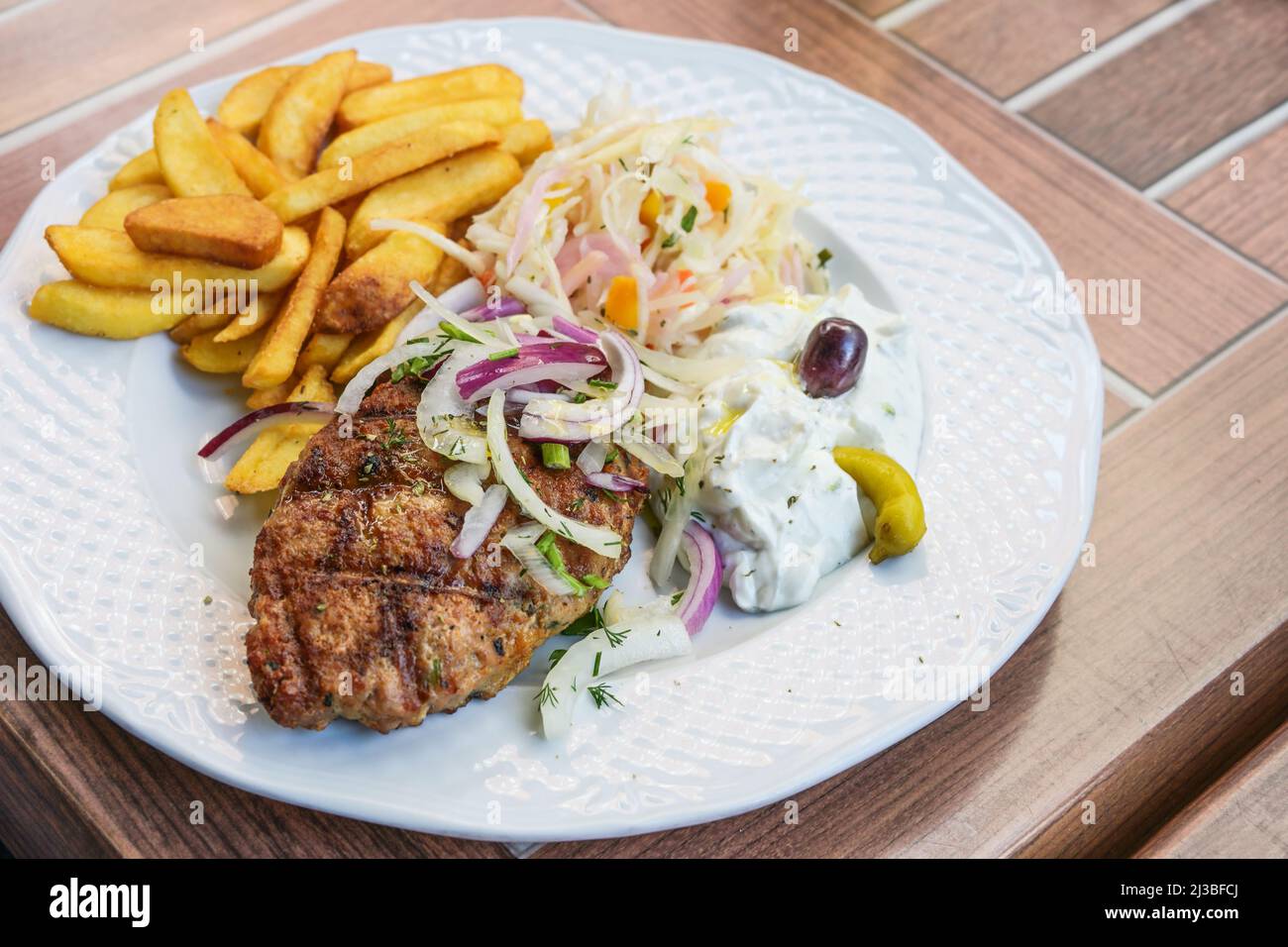 Bifteki, bistecca di carne macinata greca ripiena di formaggio feta, servita con patatine fritte, coleslaw, tzatziki e cipolle su un piatto bianco, ta in legno all'aperto Foto Stock