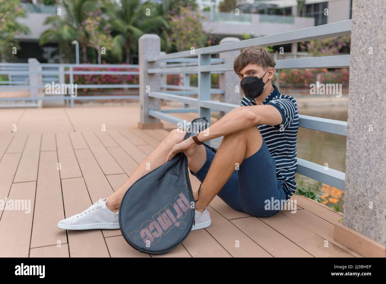 Giovane uomo caucasico con maschera nera seduta e racchetta da tennis in mano. Foto di alta qualità Foto Stock