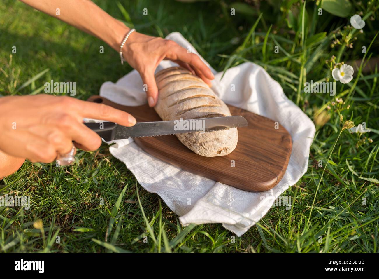 Composizione di pane fatto in casa con farina bianca su tavola di legno con tovagliolo bianco sul prato verde a mano tagliando il pane con un coltello all'aperto. Foto Stock