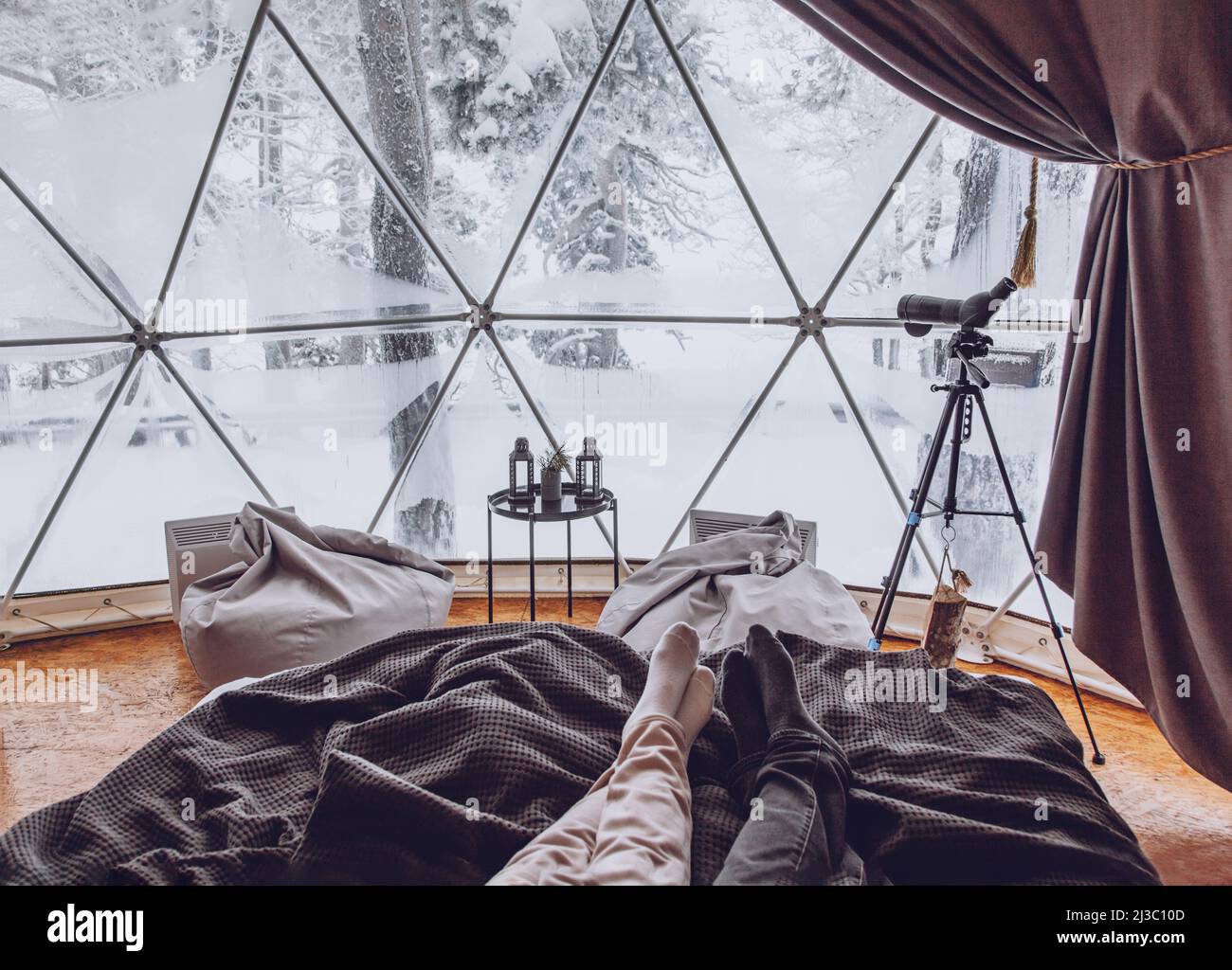 Le gambe di un uomo e di una donna si accoppiano a letto sullo sfondo di una foresta innevata in un campeggio a cupola con sedie a sacco e un telescopio su un treppiede Foto Stock
