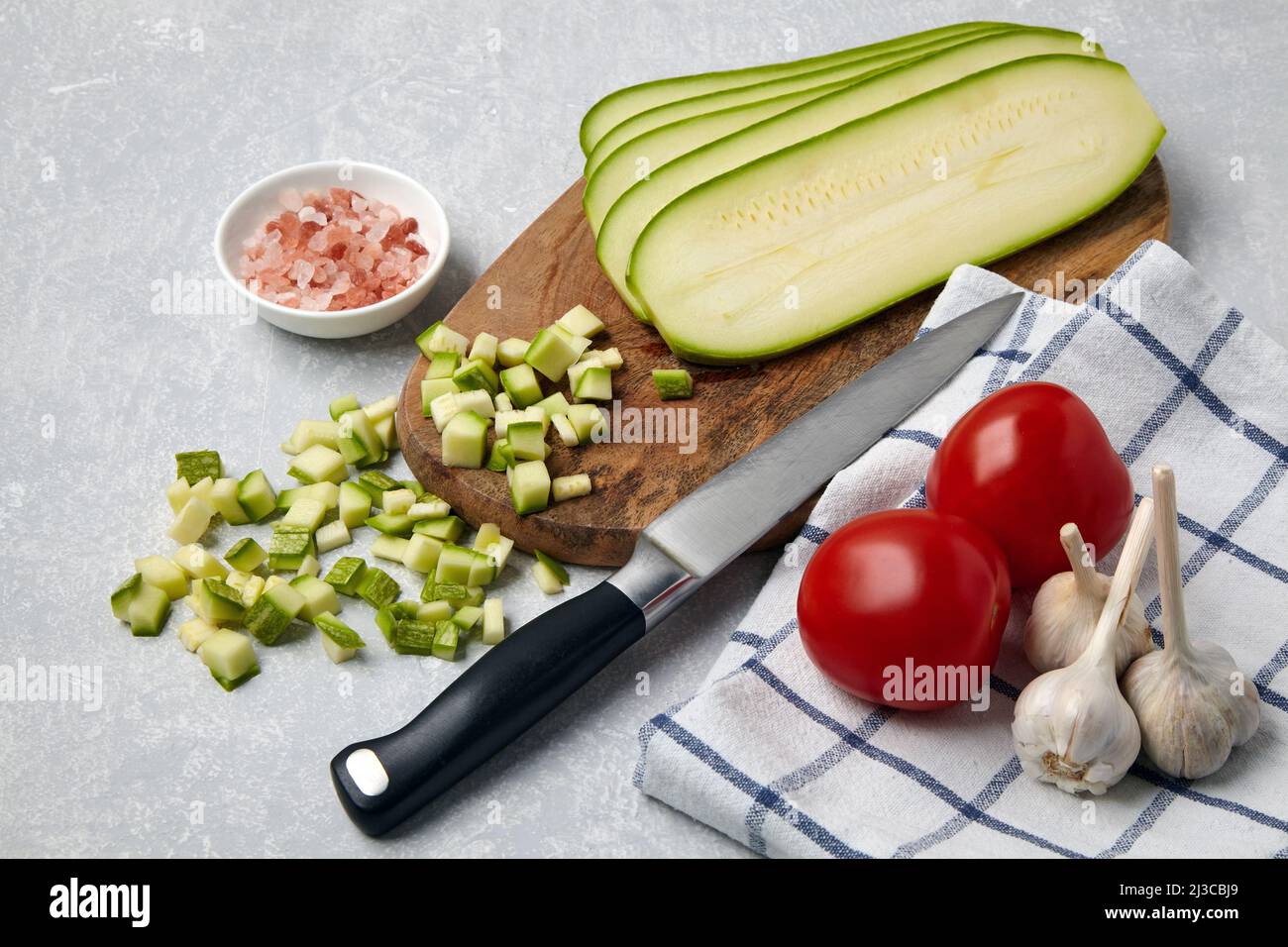 Affettato le verdure zucchine su un tagliere di legno, pomodori, aglio, sale rosa e tovagliolo su un tavolo chiaro di cemento Foto Stock