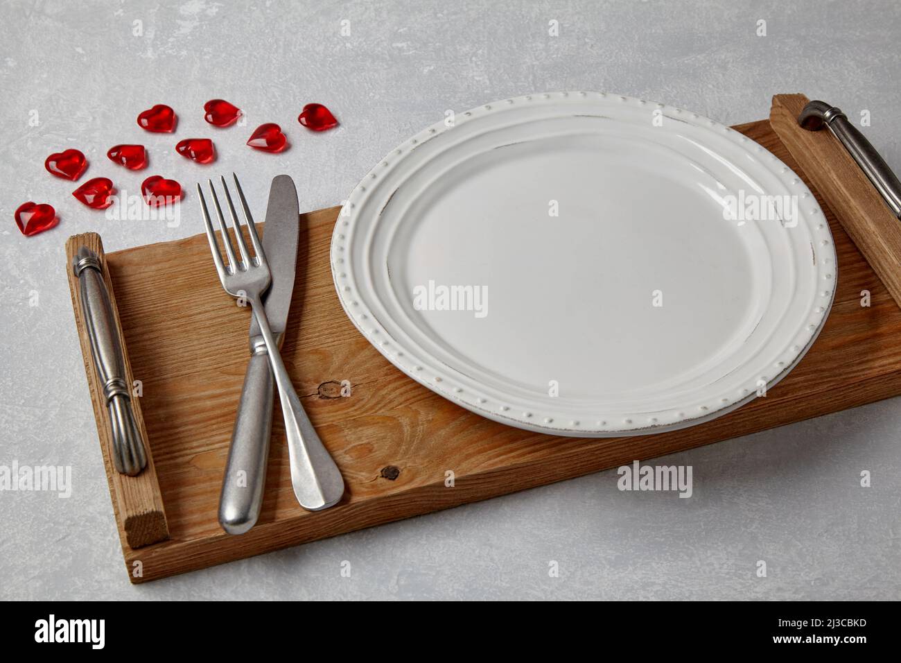 Un piatto bianco vuoto con posate su un vassoio di legno e cuori di caramello rosso su un tavolo di cemento leggero. Servizio romantico Foto Stock