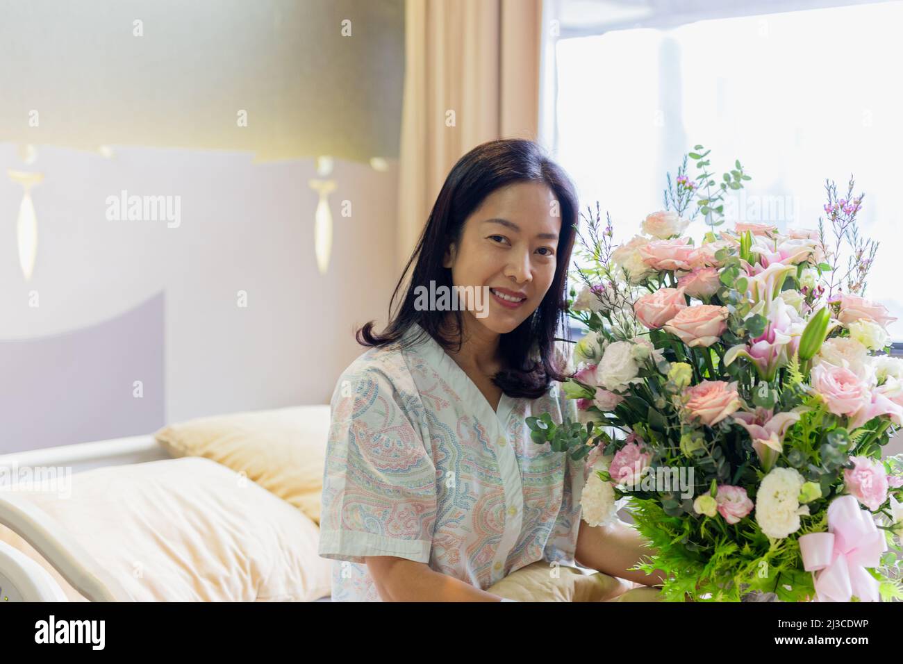 Paziente donna sorridente e con un bouquet di fiori seduto sul letto d'ospedale. Foto Stock