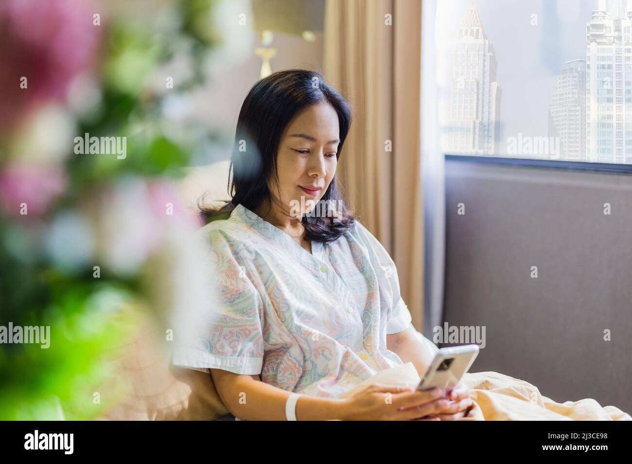 Paziente asiatico sorridente mentre si siede a letto facendo videochiamata sul cellulare in ospedale reparto. Foto Stock