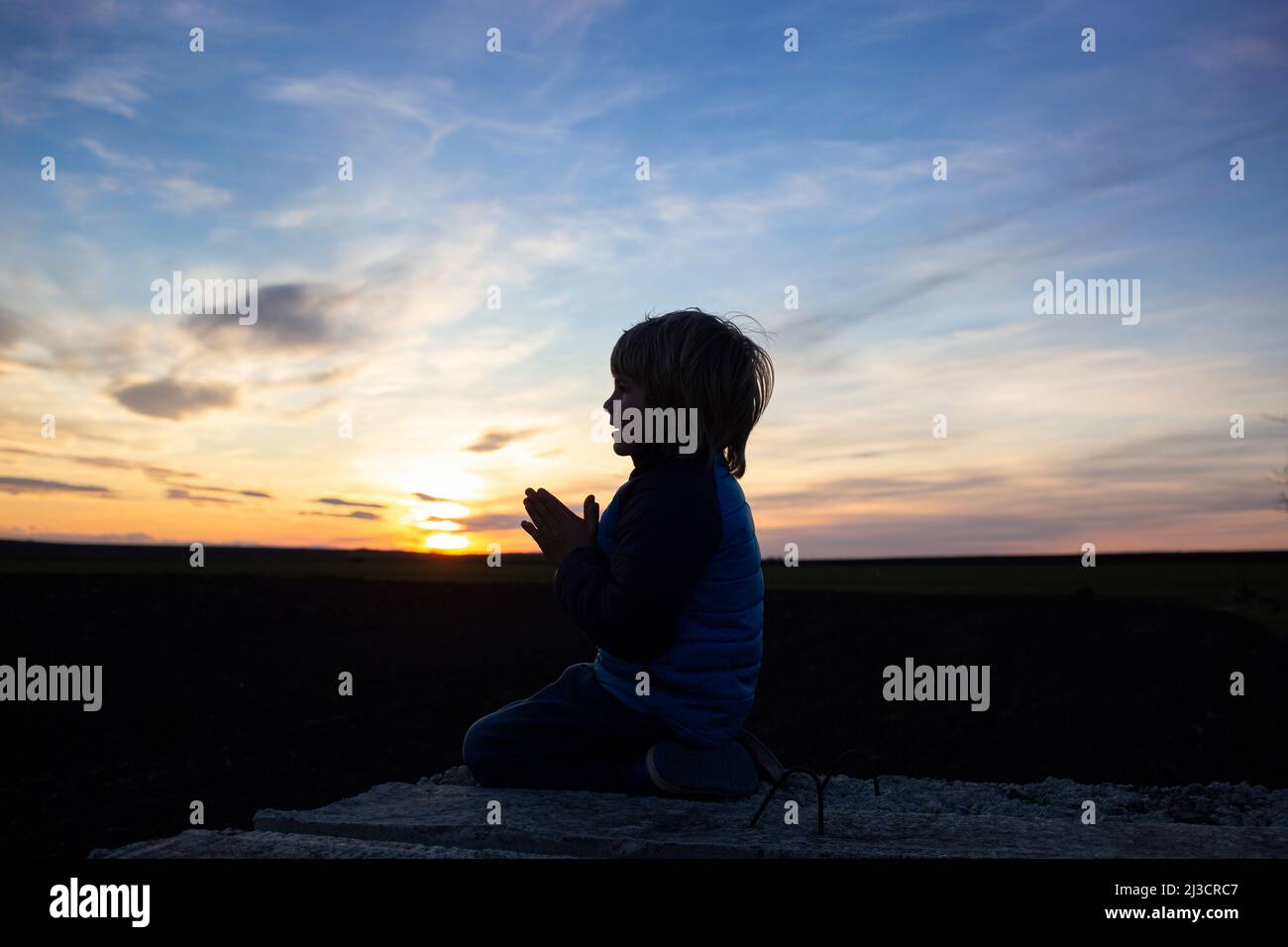 Silhouette di un ragazzo seduto sulle ginocchia, le mani piegate in preghiera, voltandosi verso Dio al tramonto. Bambini contro la guerra, il bambino chiede la pace per il suo hom Foto Stock