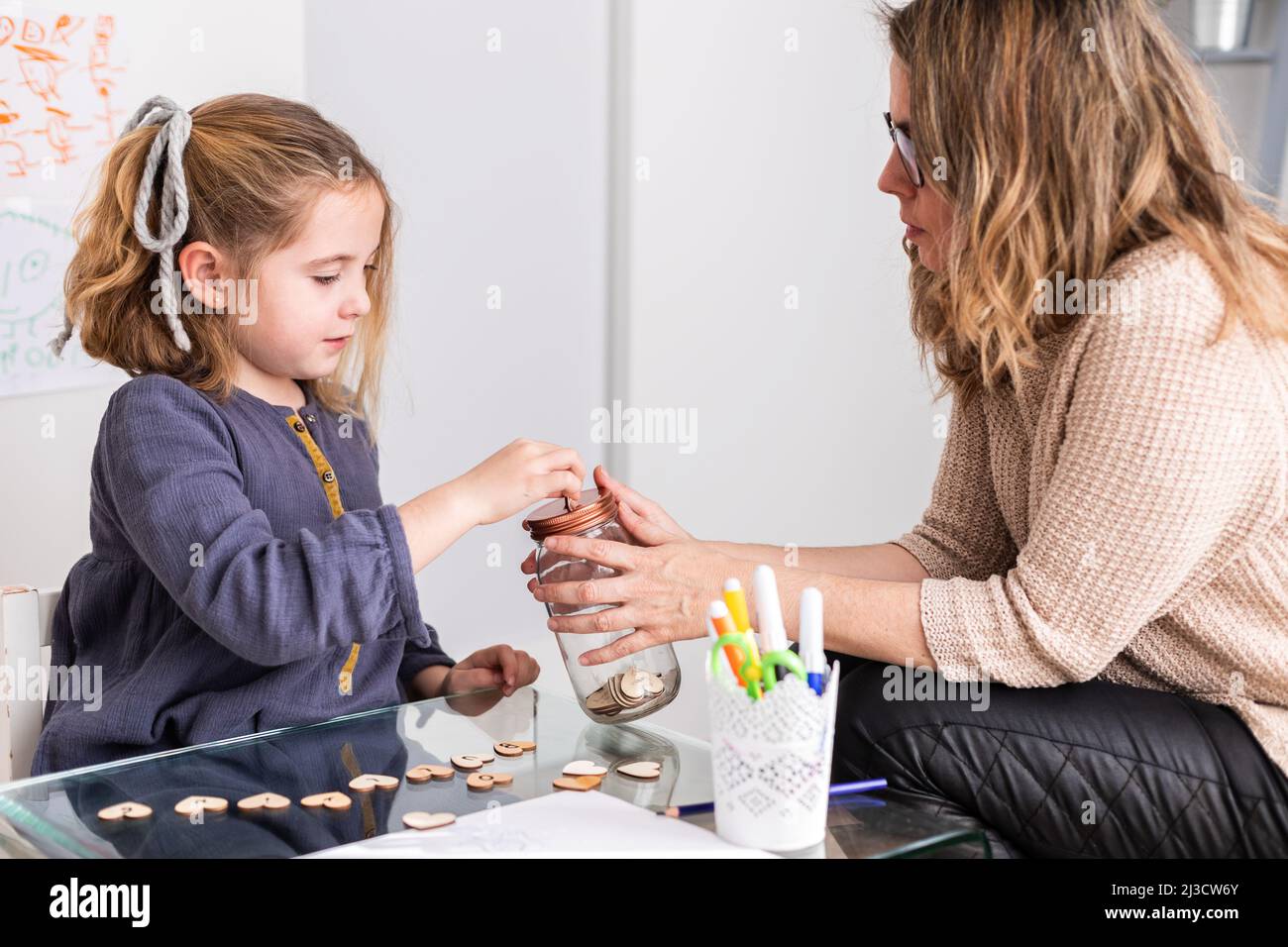 Ragazza carina che fa il desiderio con vaso di vetro piccoli cuori di legno mentre si siede vicino psicologo femminile Foto Stock