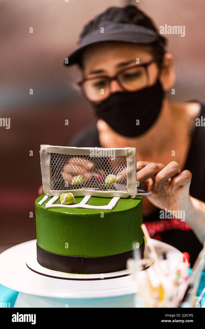 Fuoco morbido di Donna in maschera viso disposizione palle da tennis dolce vicino alla rete sulla torta fondante durante il lavoro in panetteria Foto Stock