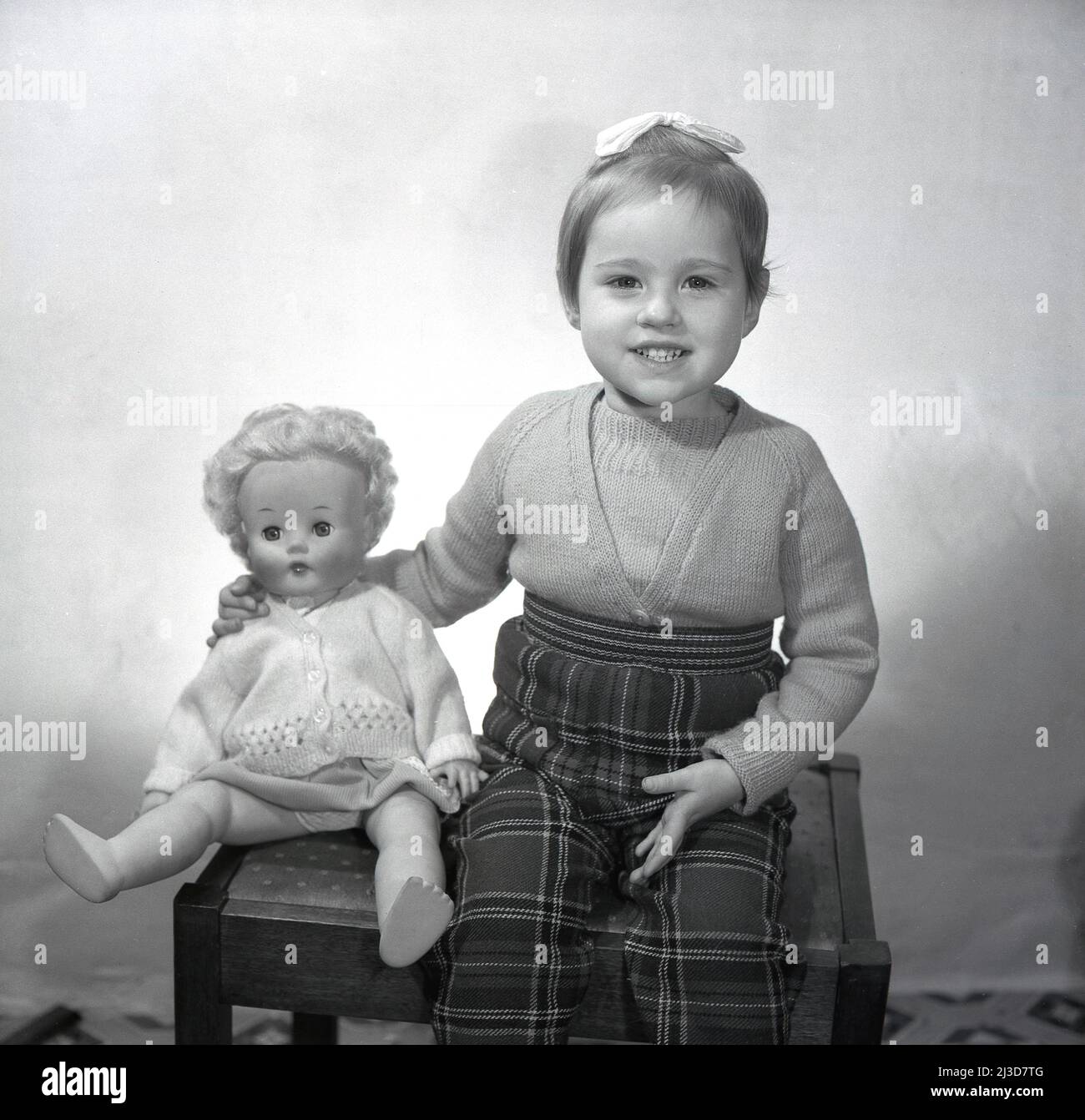 1961, storica, una bambina, con un arco nei capelli, seduta su uno sgabello per la sua foto con la sua bambola, Stockport, Manchester, Inghilterra, Regno Unito. Foto Stock