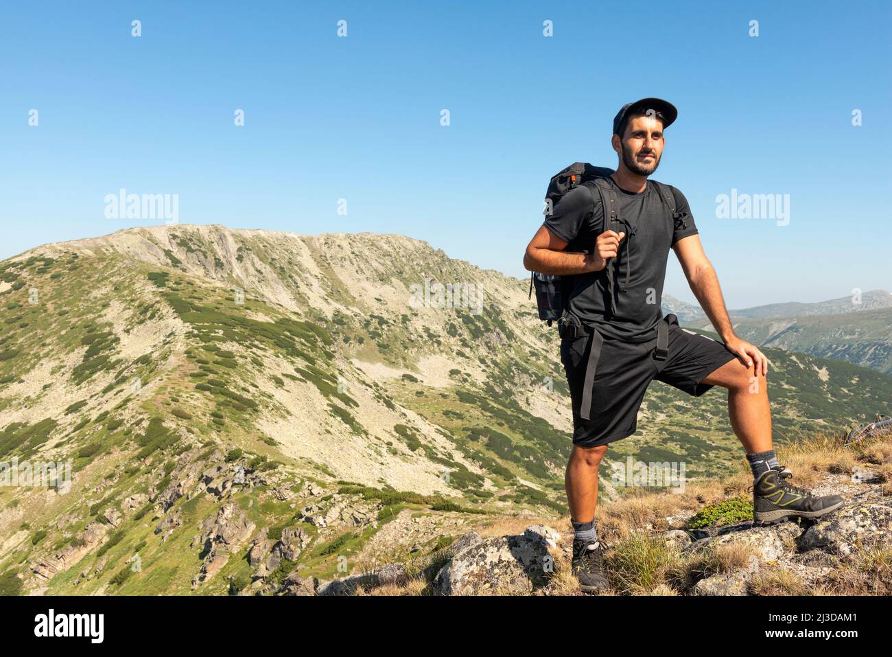 Escursionista maschile in vestito nero in posa per foto nel Parco Nazionale di Rila e Riserva Naturale, Bulgaria, Balcani. Rilets picco a 2713m sullo sfondo. Foto Stock