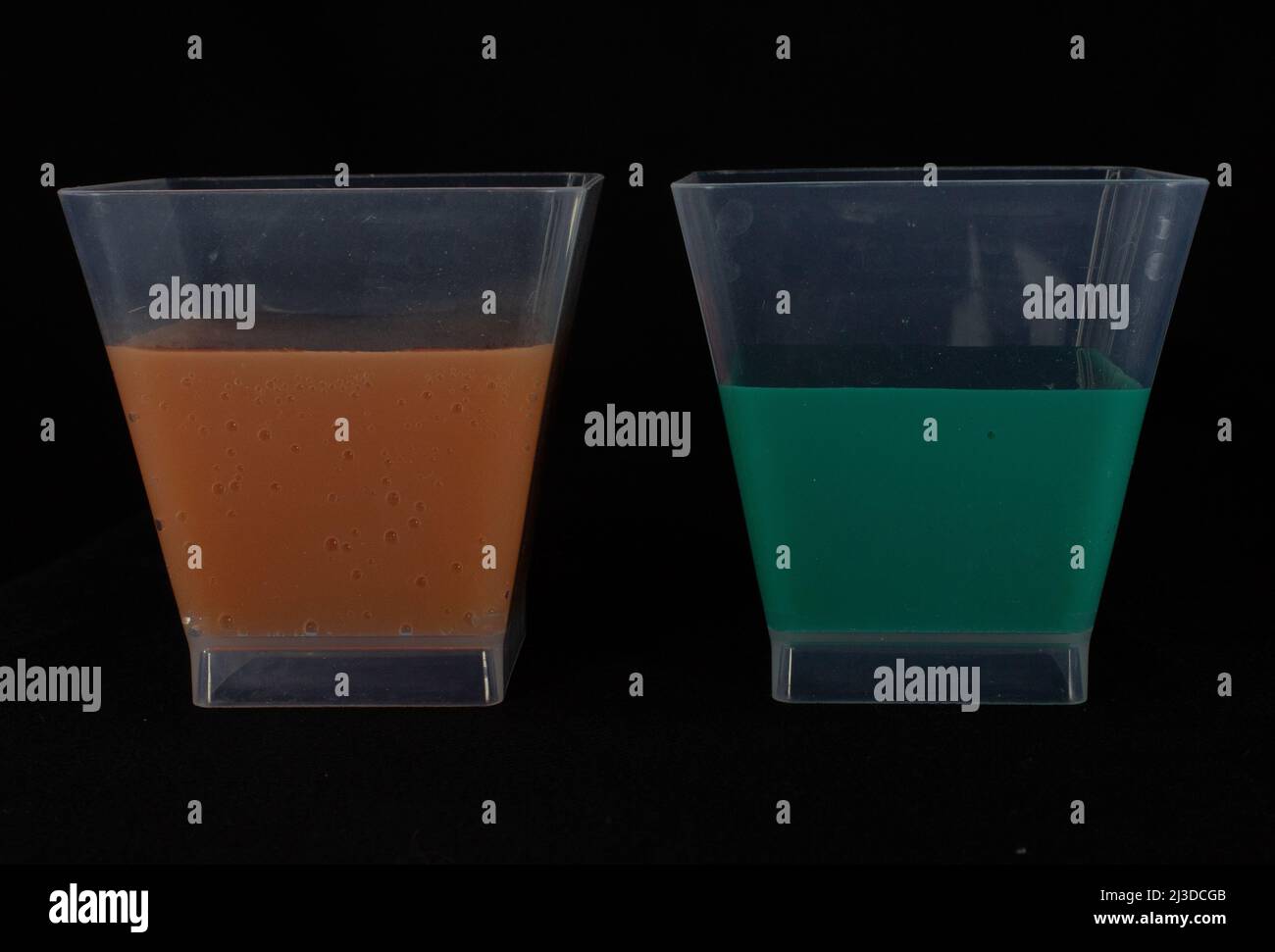 primo piano di due tazze di plastica metà piene di liquido verde e arancione isolato su sfondo nero Foto Stock