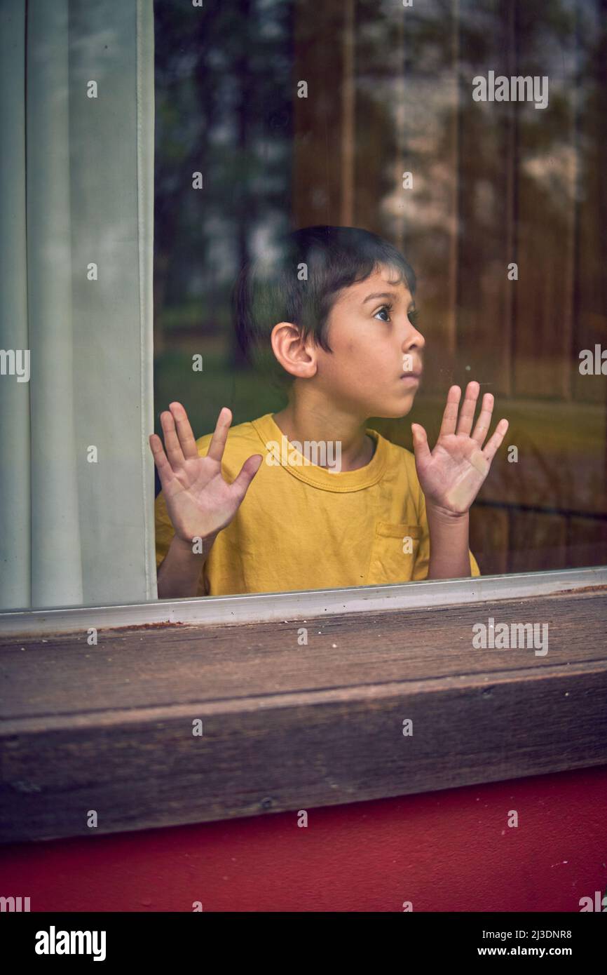 ragazzino latino solitario appoggiato le mani dietro un vetro della finestra, bloccato in una capanna nel mezzo della foresta senza uscita. Verticale Foto Stock