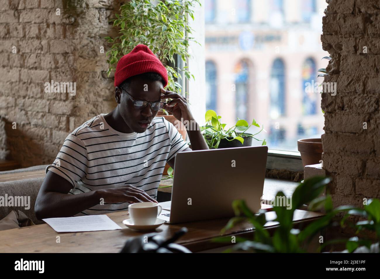 Il giovane e pensivo uomo freelance afroamericano in occhiali a distanza lavorando su un portatile in un bar Foto Stock
