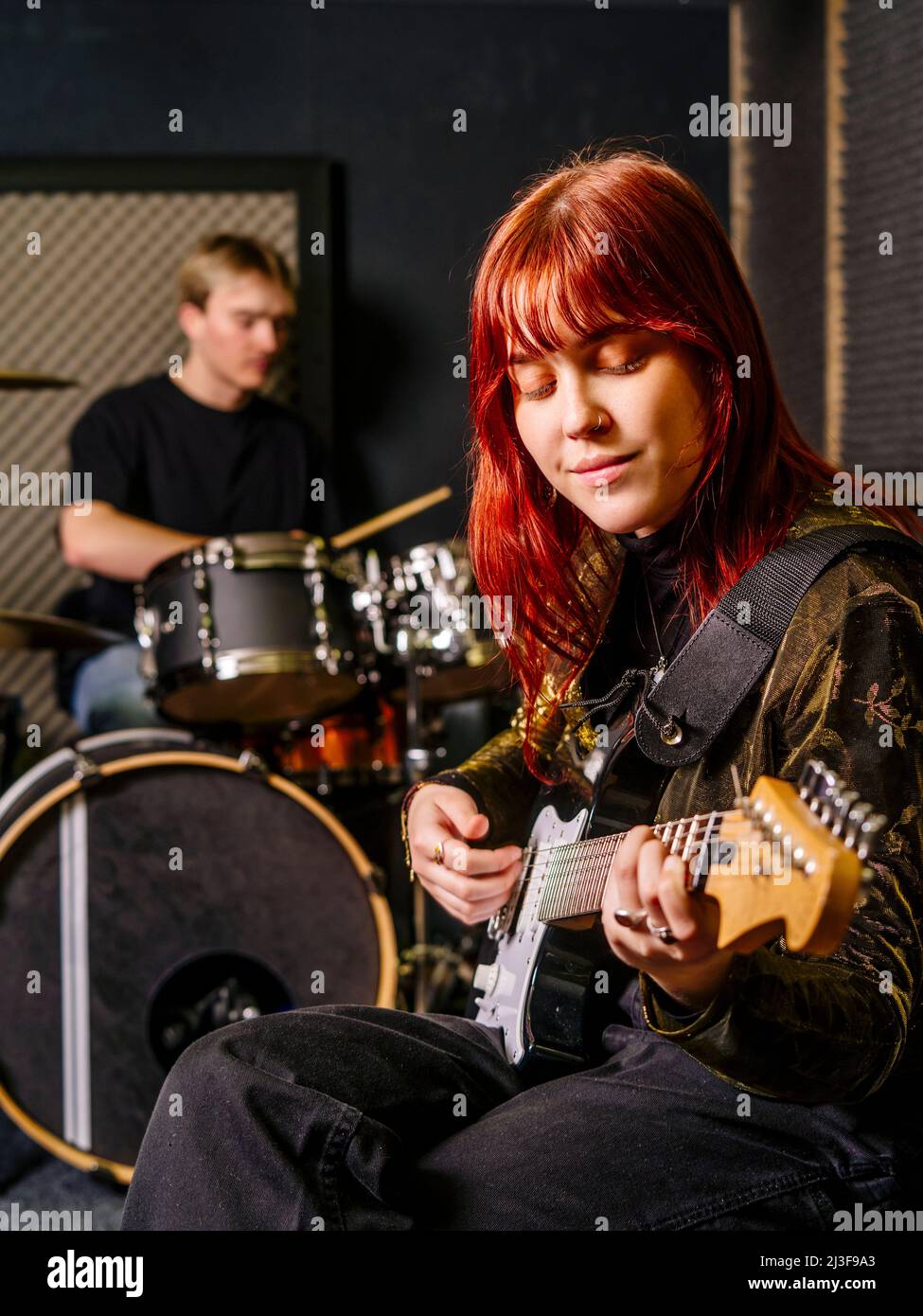 Giovane donna rossa che suona chitarra elettrica e un batterista maschile in uno studio di registrazione Foto Stock