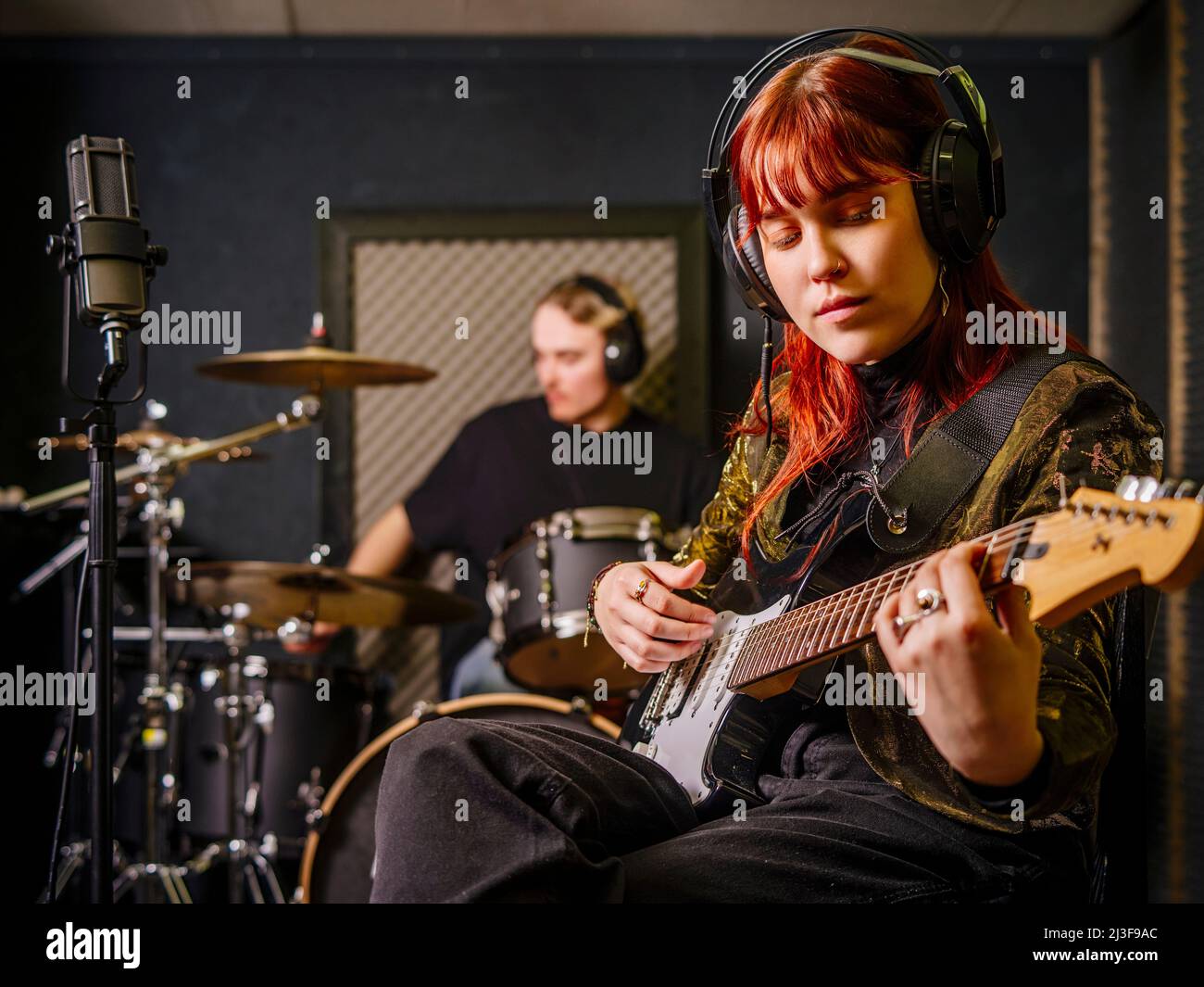 Giovane donna rossa che suona chitarra elettrica e un batterista maschile in uno studio di registrazione. Foto Stock