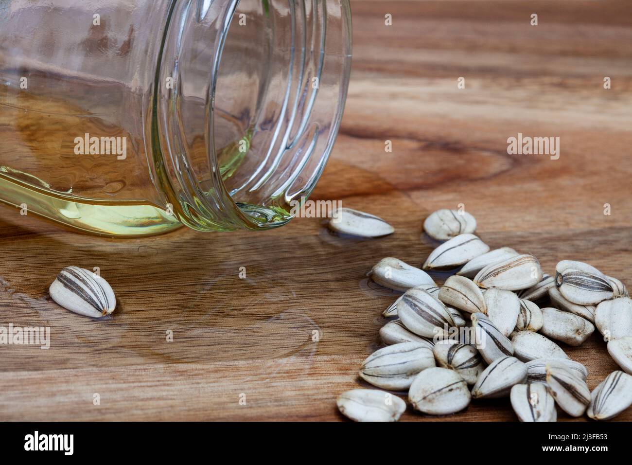Olio di girasole fuoriuscito dal vaso trasparente semplice su una superficie di legno con semi di girasole Foto Stock
