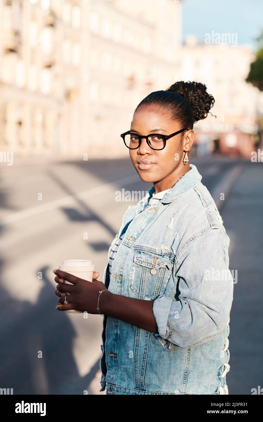 Ritratto di contenuto giovane donna nera in occhiali con giacca in denim che tiene il caffè takeout contro il paesaggio urbano Foto Stock