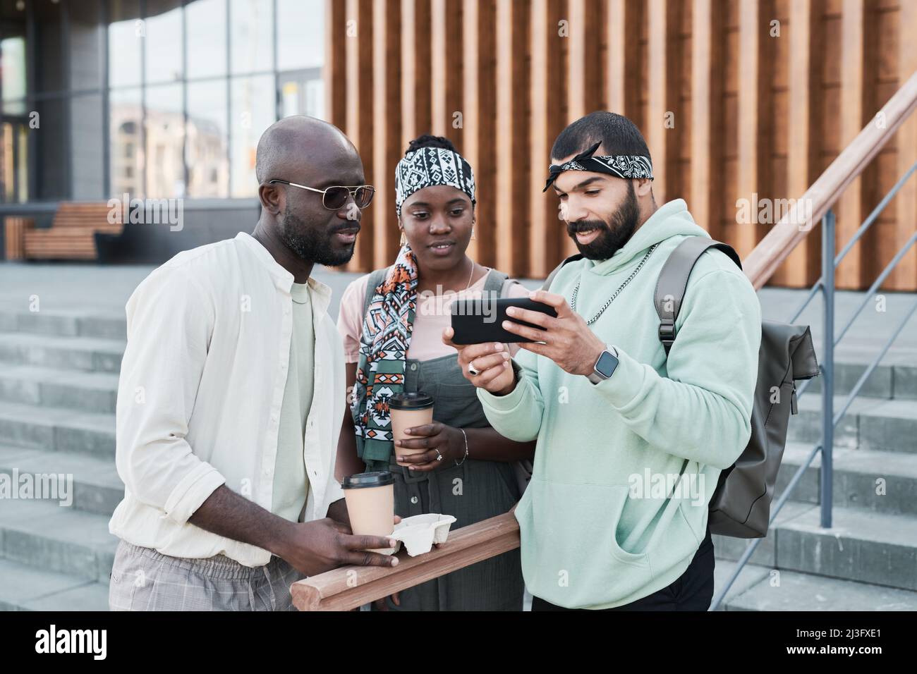 Uomo portico medio-orientale con borsa a mano che mostra foto al telefono agli amici con caffè da asporto Foto Stock