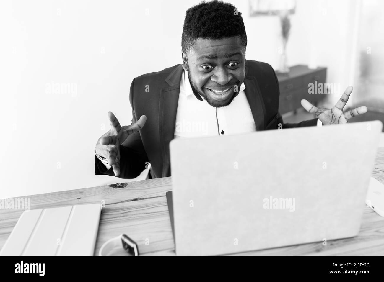 Uomo emozionale che lavora con un notebook giallo a casa o in ufficio moderno. Uomo d'affari afro-americano concentrato in abito blu, tenere il telefono vicino alla scrivania e. Foto Stock