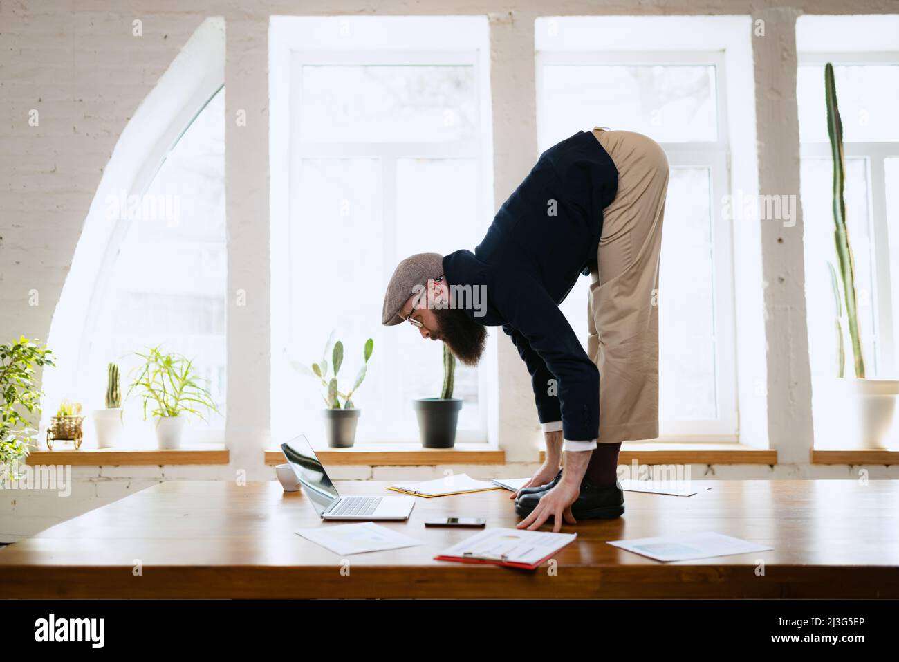 Plasticità. Giovane uomo barbuto, impiegato di ufficio che si diverte, facendo yoga su tavola di legno in ufficio moderno. Concetto di business, stile di vita sano, sport Foto Stock