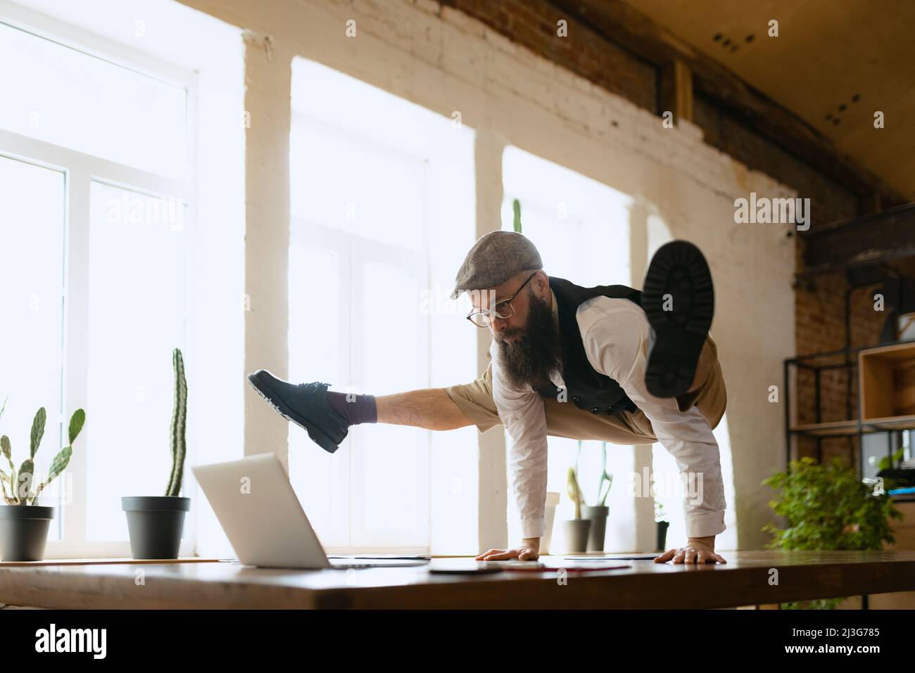 Plasticità. Giovane uomo barbuto, impiegato di ufficio che si diverte, facendo yoga su tavola di legno in ufficio moderno. Concetto di business, stile di vita sano, sport Foto Stock
