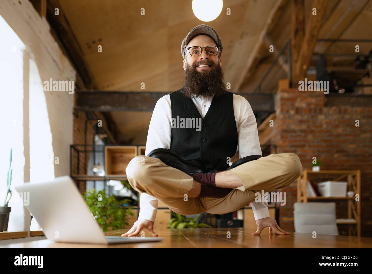 Emotivo impiegato ufficio, giovane uomo che fa yoga su tavola in legno in ufficio moderno al lavoro con gadget. Concetto di business, stile di vita sano, sport Foto Stock