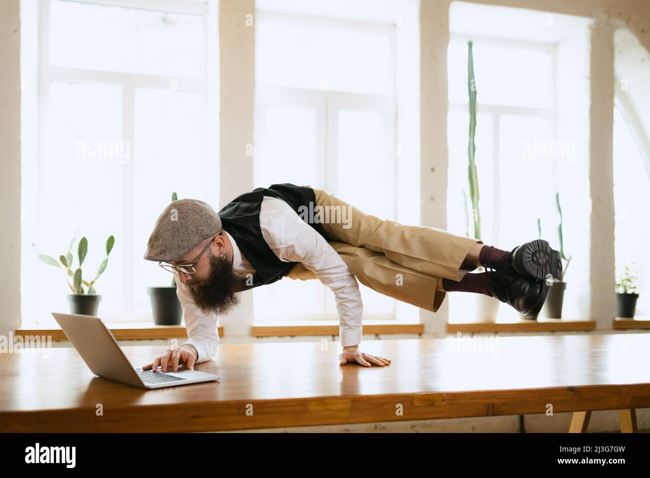 Emotivo impiegato ufficio, giovane uomo che fa yoga su tavola in legno in ufficio moderno al lavoro con gadget. Concetto di business, stile di vita sano, sport Foto Stock