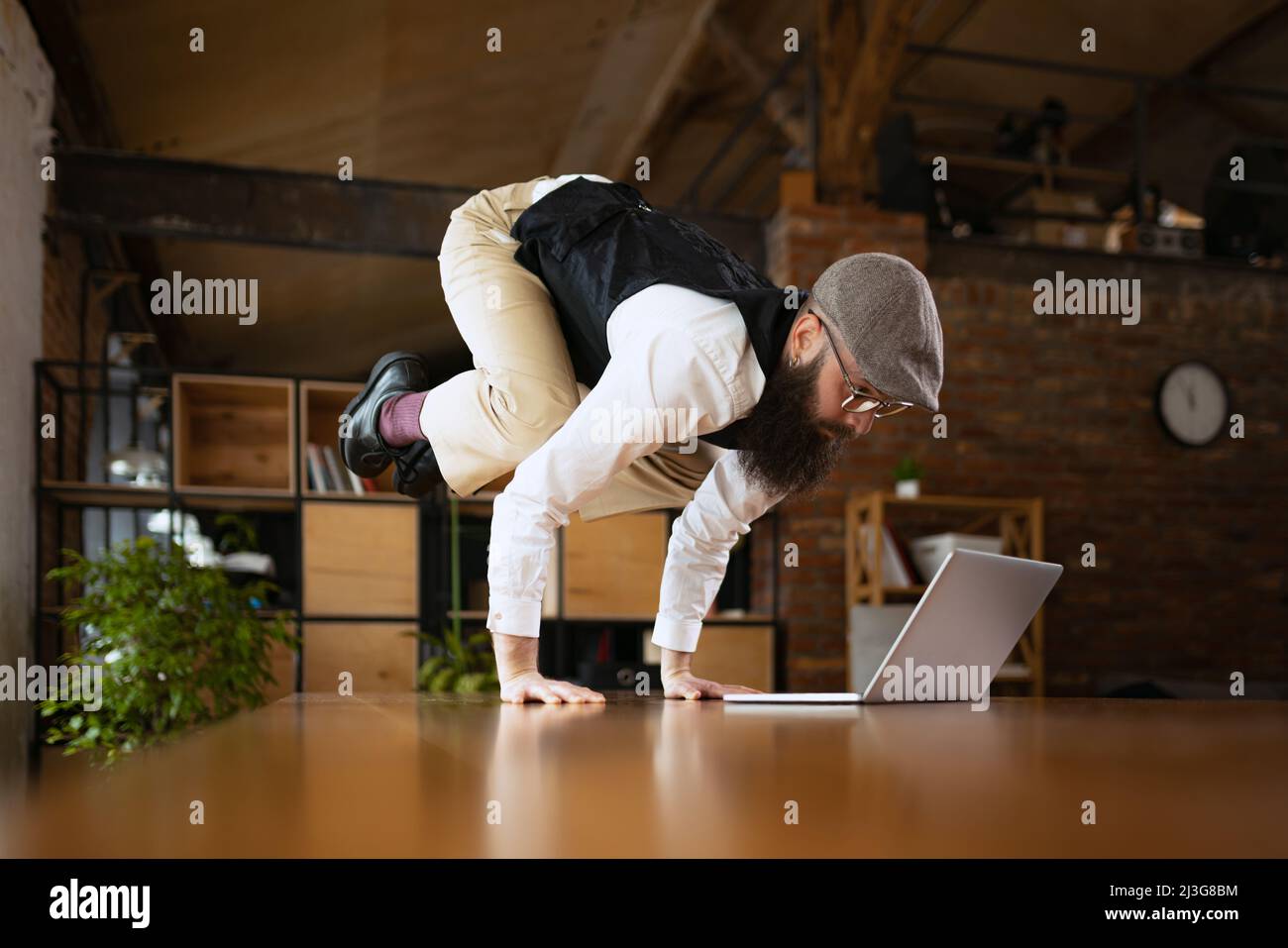 Riunione on line. Giovane uomo barbuto, impiegato di ufficio che si diverte, facendo yoga su tavola di legno in ufficio moderno al tempo di lavoro con i gadget. Concetto di Foto Stock