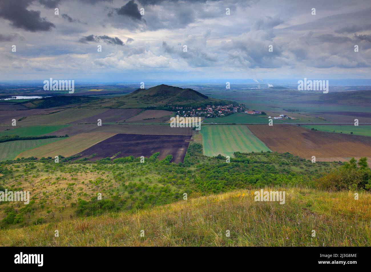 Rana Hill, nella montagna di Ceske Stredohori, Repubblica Ceca. Collina con le nuvole. Bellissimo paesaggio mattutino. Villaggio sotto la collina. Paesaggio con campi Foto Stock