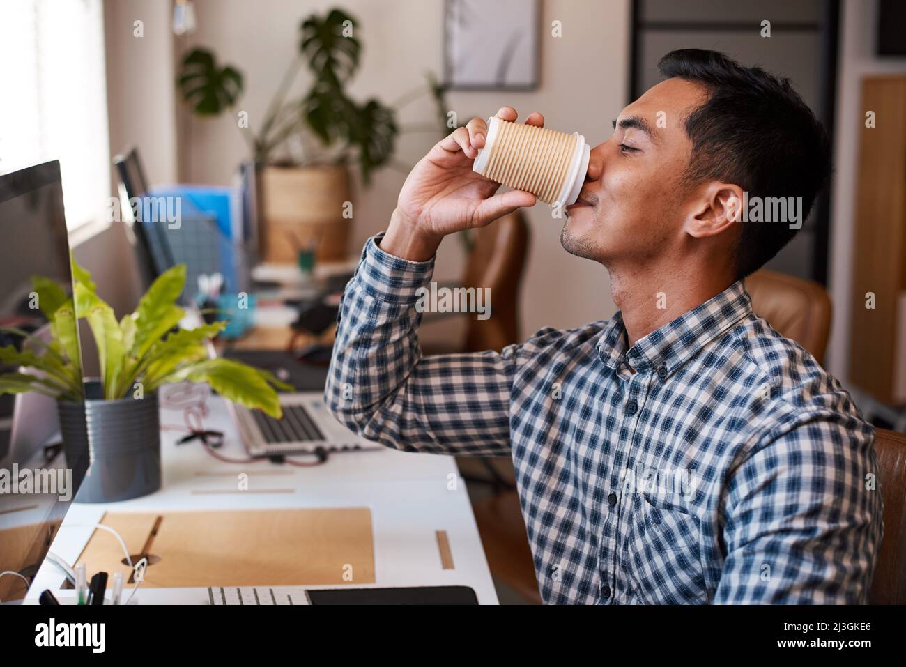 Un giovane uomo d'affari asiatico beve caffè alla sua scrivania in un ufficio vuoto Foto Stock