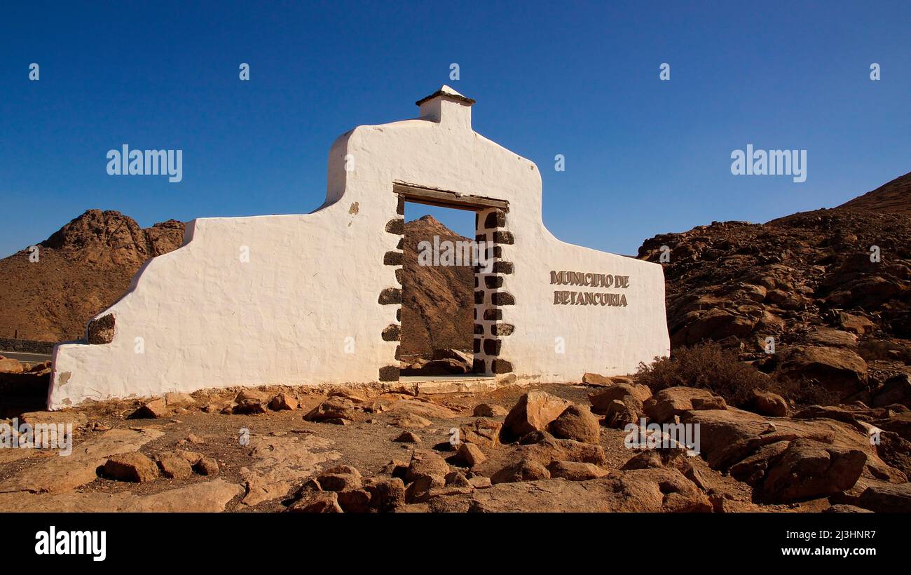 Spagna, Isole Canarie, Fuerteventura, punto di vista, Mirador de la Penitas, arco in pietra bianca muratura che indica il comune di Bethancuria, in paesaggio roccioso, azzurro cielo e nuvoloso Foto Stock