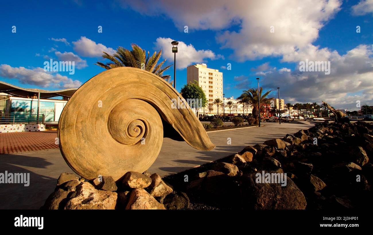 Spagna, Isole Canarie, Fuerteventura, capitale, Puerto del Rosario, installazione artistica, lumaca o croissant in cemento, in piedi, passeggiata del porto, edificio sullo sfondo, azzurro cielo, nuvole bianche Foto Stock