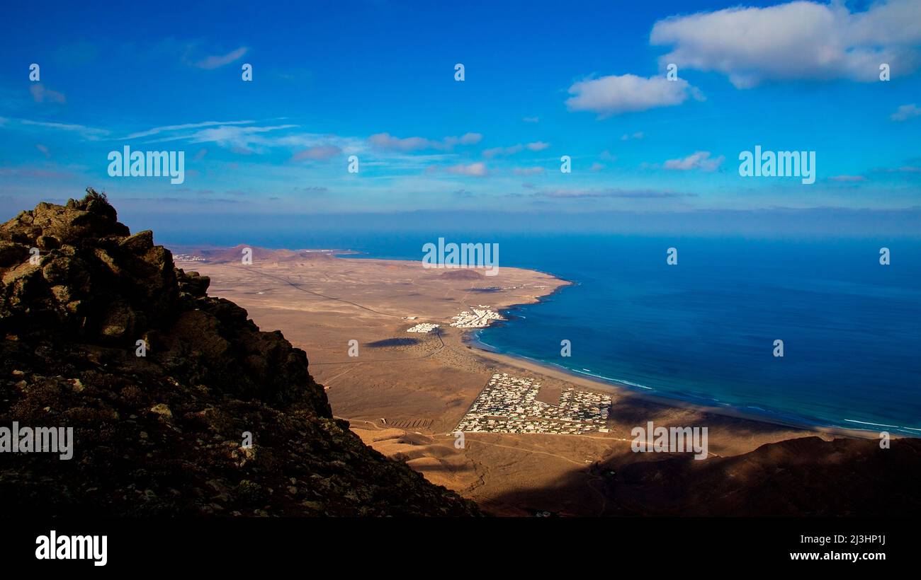 Isole Canarie, Lanzarote, isola vulcanica, costa nord-occidentale, eremo, chiesa, Ermita de las Nieves, cielo blu, grandangolo colpo, vista dall'Ermita fino alla costa occidentale, nelle rocce in primo piano, sotto si può vedere un insediamento turistico bianco, cielo blu con nuvole bianche, mare blu e verde Foto Stock