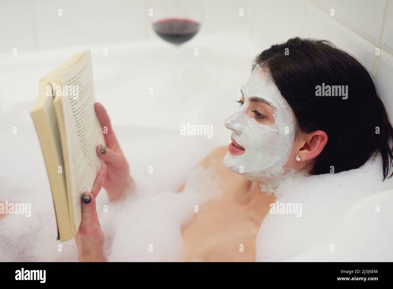 Trattandosi ad un certo tempo nella vasca. Scatto corto di una giovane donna che si rilassa nella vasca da bagno con un libro e un bicchiere di vino. Foto Stock