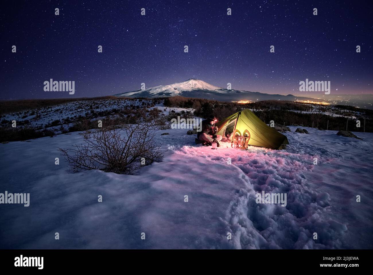 Uomo che riposa vicino alla sua tenda illuminata sulla neve di notte sotto il cielo stellato, sullo sfondo il Monte Etna dal Parco dei Nebrodi, in Sicilia Foto Stock