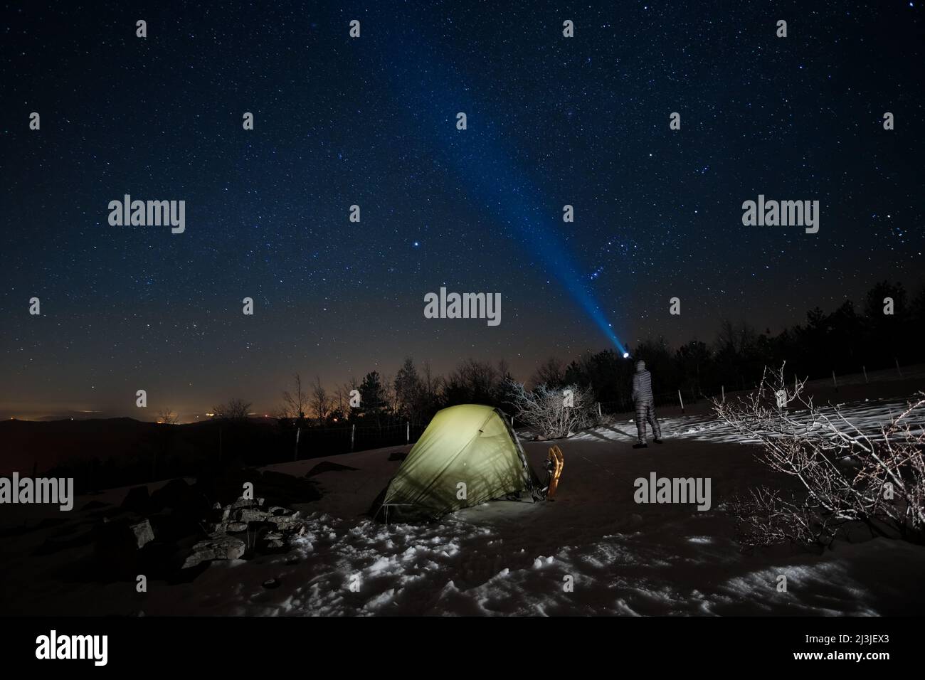 Uomo in piedi vicino alla tenda illuminata e puntando la torcia blu al cielo stellato in inverno Nebrodi Park, Sicilia Foto Stock