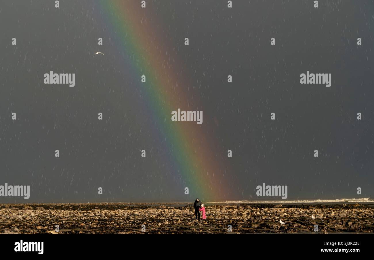 Padre e figlia si levano in piedi piccoli contro un vasto arcobaleno in un cielo scuro durante una pioggia; Whitburn, Tyne e Wear, Inghilterra Foto Stock