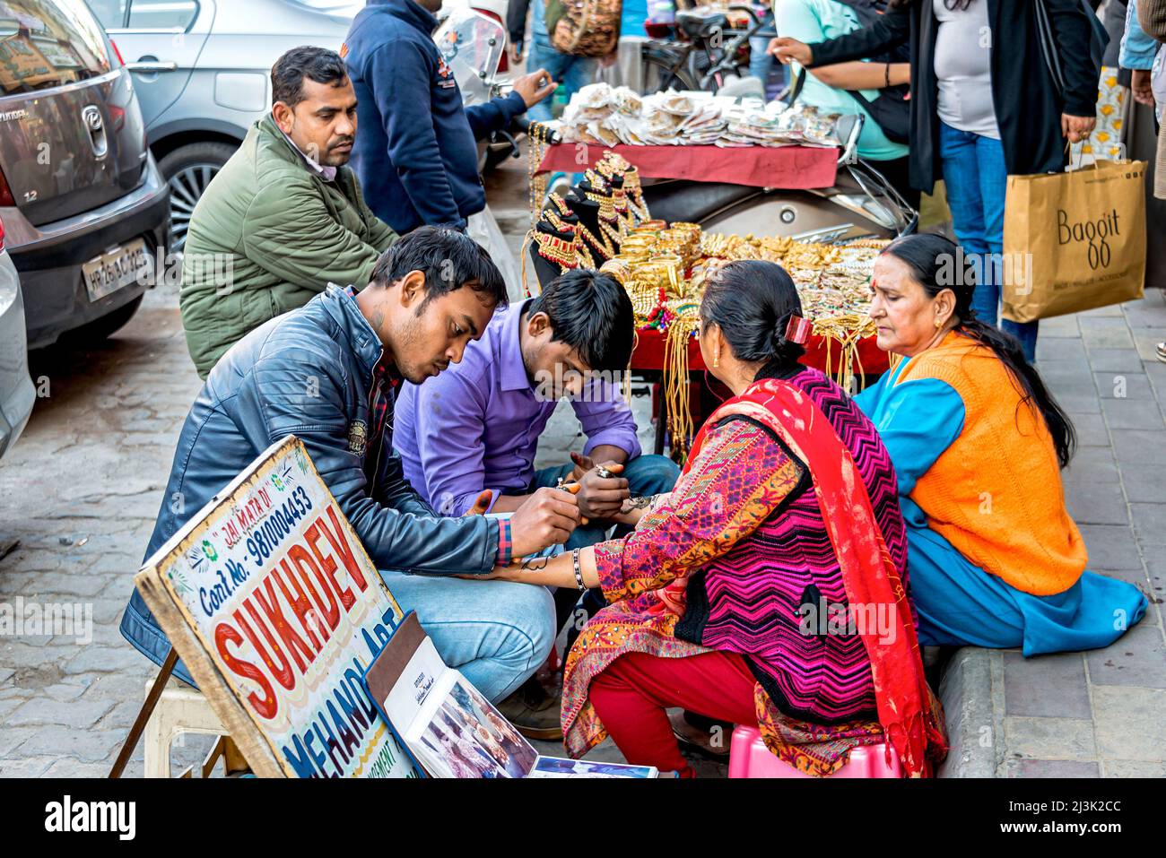 Donna che ottiene tatuaggi henné (Mehndi Art) sulle mani mentre si siede su una strada cittadina accanto a un fornitore di gioielli; Amritsar, Punjab, India Foto Stock