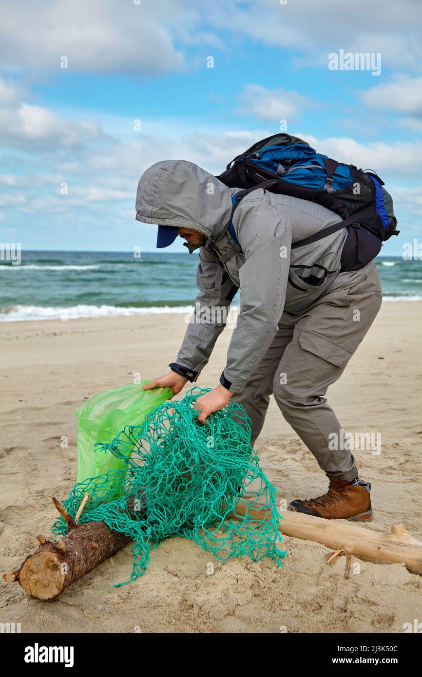 Il volontario maschile in tempo ventoso raccoglie rifiuti gettato da una tempesta su una spiaggia di sabbia. Il problema dell'inquinamento degli oceani e degli ambientani del mondo Foto Stock