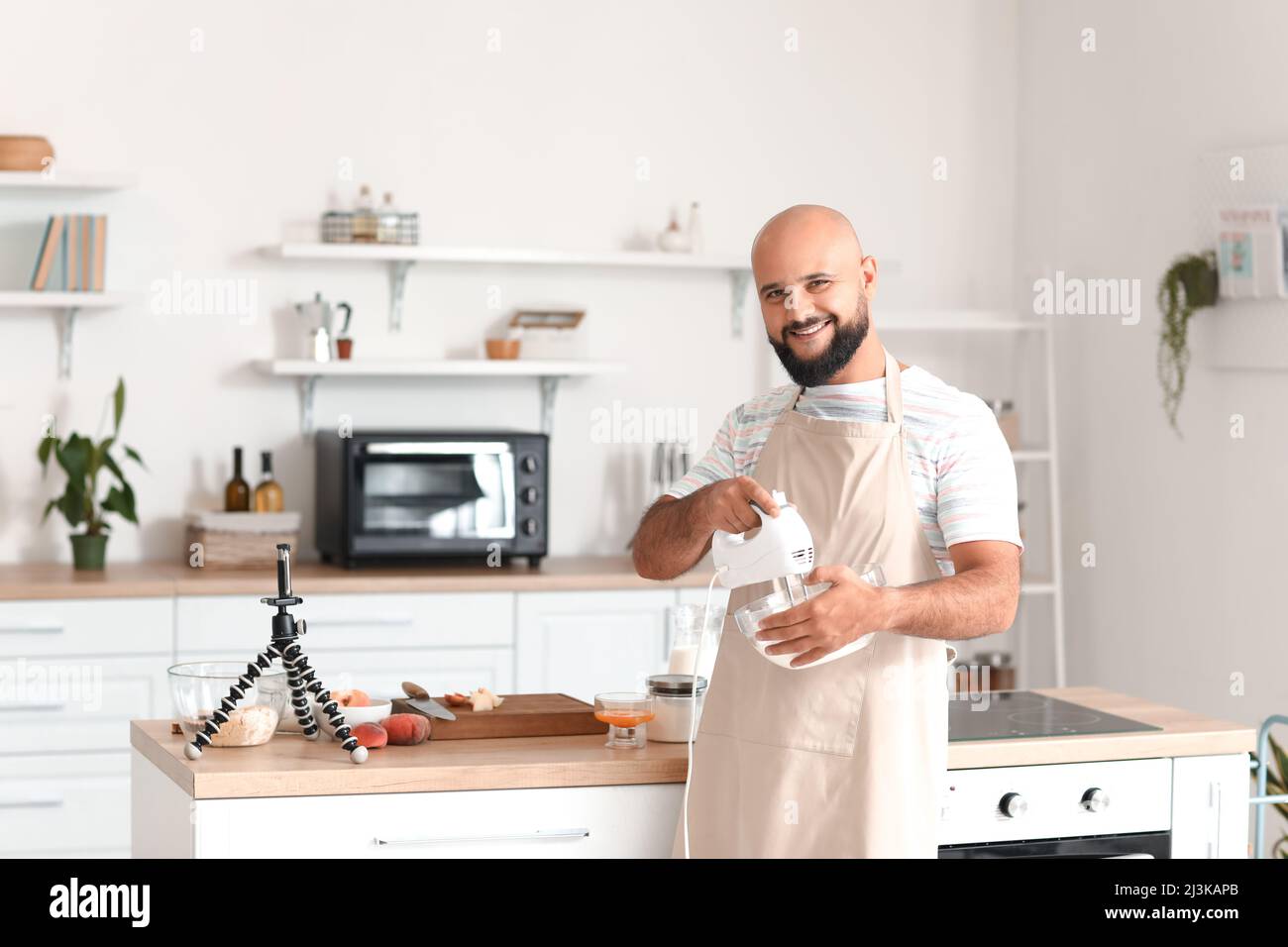 Bell'uomo che frusta l'albume d'uovo con il mixer mentre segue il video tutorial di cucina in cucina Foto Stock