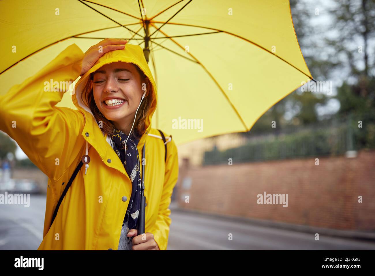 Primo piano di una giovane donna allegra con un impermeabile giallo e un ombrello che è di buon umore mentre si cammina per la città in una giornata piovosa in modo rilassato Foto Stock