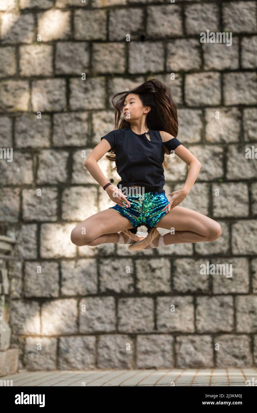 Ragazza che balla con scarpe da balletto e in posa a mezz'aria contro un muro di pietra in un parco pubblico; Hong Kong, Cina Foto Stock